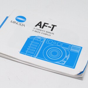 Puede incluir: Un manual de usuario de la cámara Minolta AF-T de color blanco con una ilustración azul de la cámara en la portada. El texto en la portada dice "AF-T Owner's Manual Mode d'Emploi".