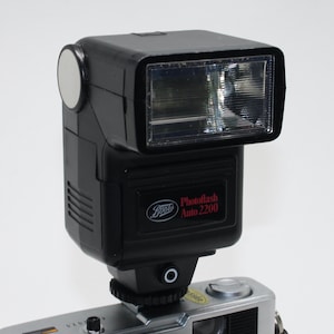 Könnte beinhalten: Ein schwarzes Boots Photoflash Auto 2200 Blitzgerät an einer silbernen Kamera befestigt. Der Blitz hat einen rechteckigen Kopf mit einer klaren Linse und einen runden silbernen Reflektor. Die Kamera ist ein Trip 35 Modell.