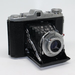 Puede incluir: Una cámara plegable vintage en blanco y negro con el texto "Wefamy G" en la parte superior. La lente es plateada con texto negro y un indicador rojo. La cámara está abierta, revelando el fuelle.