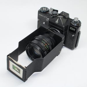 Puede incluir: Una cámara Zenit EM negra con una lente y un visor. La cámara tiene un dial plateado y la palabra "ZENIT" está impresa en la parte superior. La lente tiene una serie de números y colores. El visor tiene una imagen pequeña.