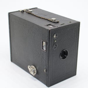 May include: A vintage black box camera with a leather handle and a textured surface. The camera features a lens, viewfinder, and various dials and buttons. This classic camera is an example of early photography equipment.