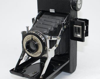 May include: A vintage black Kershaw Penguin Eight-20 camera with a leather strap. The camera has a silver lens and a black bellows. The camera is open, revealing the lens and internal components. The text "Kershaw Eight-20" and "Penguin" are visible on the lens.