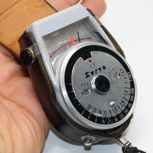Puede incluir: Un fotómetro vintage ST132 con esfera plateada y marcas en blanco y negro. Está alojado en una funda de cuero negro con una correa de cuero marrón. El dial incluye configuraciones ASA, DIN y F-STOP.