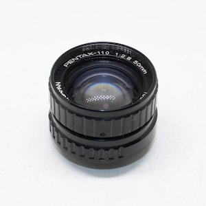 Puede incluir: Un objetivo de cámara negro Pentax-110 de 50 mm. El objetivo tiene un anillo de enfoque estriado y un elemento de vidrio transparente. El texto "PENTAX-110" y "50mm" es visible en el objetivo. Fabricado en Japón.