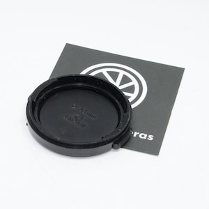 May include: A black camera lens cap with the text "28-80 mm" and "JAPAN" is displayed on a white background. The lens cap is circular with a ridged edge. A black and white card with a logo is in the background.