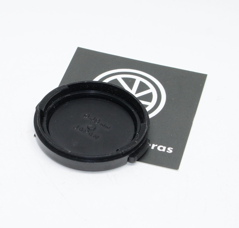 May include: A black camera lens cap with the text "28-80 mm" and "JAPAN" is displayed on a white background. The lens cap is circular with a ridged edge. A black and white card with a logo is in the background.