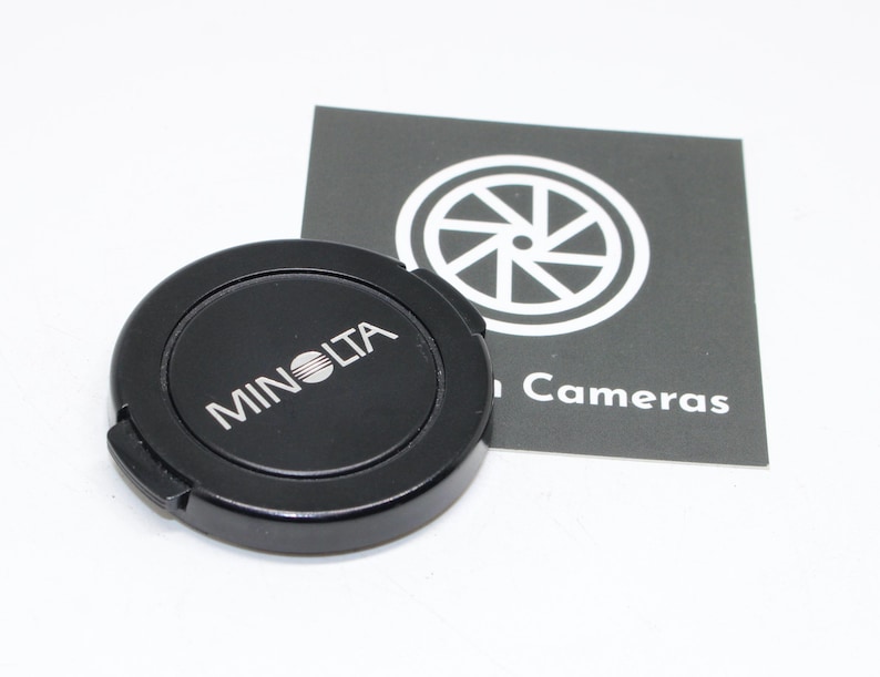 May include: A black Minolta lens cap with white lettering and logo. A black and white card with a camera lens graphic and the word "Cameras" is next to the lens cap. The lens cap is round and has a small tab for easy removal.