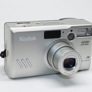 以下が含まれることがあります： 35-80mmレンズを搭載した、シルバーのKodak KV3580 Date Backカメラ。内蔵フラッシュと黒いリストストラップが付いています。前面にはKodakのロゴが見えます。