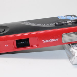 May include: A black and red SupaSnaps 110 film camera with a built-in flash and a black wrist strap. The camera features the text SupaSnaps, 110, and Pocket on its body. A silver foil-wrapped roll of Tiger 200 ISO color film sits beside it.