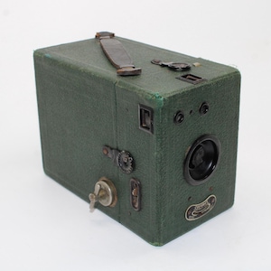 May include: A vintage box camera covered in dark green textured material with a brown leather handle on top. The front features a black circular lens, two small viewfinders, and a metal plate with the text Coronet Portrait Camera.