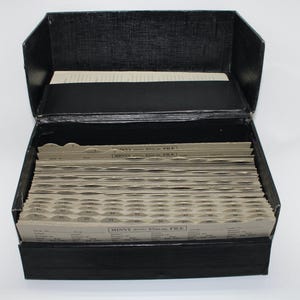 Puede incluir: Caja de archivo negra abierta con tapa con bisagras, llena de tarjetas de índice numeradas. Las tarjetas tienen un borde con muescas y están dispuestas en filas. La caja está hecha de un material texturizado.