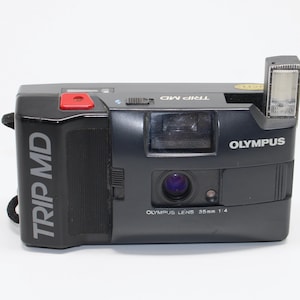 Puede incluir: Cámara de película Olympus Trip MD negra de 35 mm con flash incorporado. La cámara tiene un botón rojo y el texto "TRIP MD" está impreso en el lateral. La lente está etiquetada como "OLYMPUS LENS 35mm 1:4".