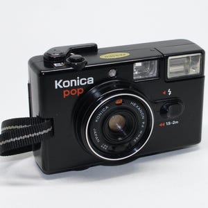 Peut inclure: Appareil photo Konica Pop noir avec une bague d'objectif argentée et une dragonne rayée noire et blanche. Le texte "Konica pop" est en blanc et rouge, et "HEXANON F4 36mm" sur l'objectif.