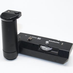 Puede incluir: Accesorio de cámara negro Olympus OM Winder 2. El winder tiene un agarre cilíndrico y una base rectangular con un dial y el logotipo de Olympus. El accesorio está diseñado para acoplarse a un cuerpo de cámara.