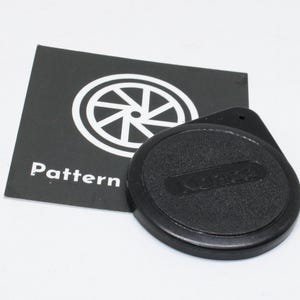 Peut inclure: Un bouchon d'objectif d'appareil photo noir avec le mot "KODAK" en relief, à côté d'une carte en noir et blanc avec le mot "Pattern" et un motif circulaire. Le bouchon d'objectif est rond et a une surface texturée.
