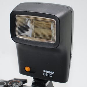 Könnte beinhalten: Ein schwarzes Prinz 880C Kamera-Blitzgerät an einem silbernen Kameragehäuse befestigt. Der Blitz hat einen rechteckigen Kopf mit einer klaren, geriffelten Linse. Auf der Vorderseite ist ein orangefarbener Knopf zu sehen. Der Text "PRINZ 880C" ist auf der Seite aufgedruckt.