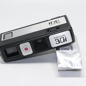 May include: A black and silver Tudormatic 301 camera with a pink flower design. The camera is sitting on a white surface.