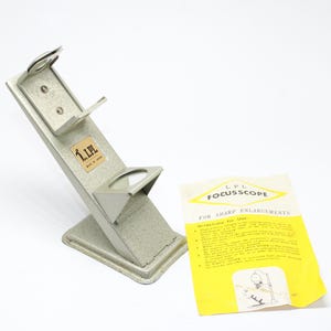 May include: A vintage LPL Focuscope for sharp enlargements. The device is made of silver-coloured metal and has a unique angled design with two circular holders. A yellow and white instruction sheet is included.