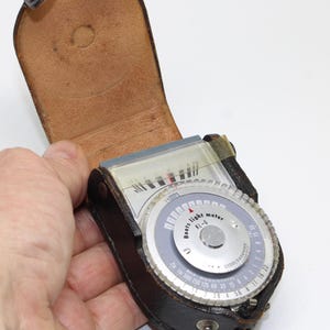 May include: A vintage Boots light meter KL-6 housed in a brown leather case with a hinged lid. The device features a circular silver dial with black numerical markings, a clear glass display window, and a needle indicator for measuring light levels.