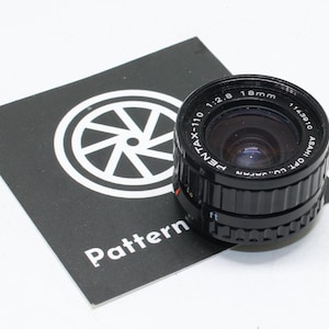 Puede incluir: Un objetivo de cámara negro Pentax-110 18mm f/2.8 con el texto "ASAHI OPT. CO., JAPAN" y un gráfico en blanco y negro sobre una tarjeta negra. El objetivo tiene aproximadamente 2,5 cm de diámetro.