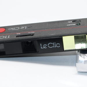 May include: A black Le Clic 110TF film camera with a red shutter button and a flash. The camera has the words "Le Clic" and "110TF" printed on it in white. A silver film pack is attached to the camera.