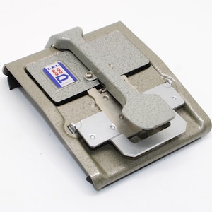 Pode incluir: Um emendador de filme LPL S-8 de metal cinza com acabamento texturizado, um braço de alavanca central e uma etiqueta retangular com detalhes em azul, branco e vermelho. O dispositivo inclui placas de metal prateado e componentes mecânicos.