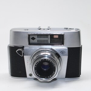 May include: A vintage silver and black Agfa Color Apotar camera. The camera has a lens with the text "COLOR APOTAR 1:2.8/45" and a viewfinder. The camera body is silver with black leatherette sides.