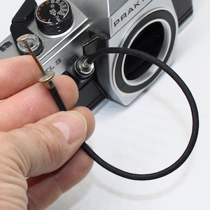 May include: A black fabric-covered mechanical shutter release cable with a silver metal plunger tip, attached to a silver and black Praktica MTL 5 film camera body. The cable forms a circular loop, and the plunger features a textured top surface.
