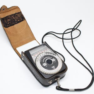 Puede incluir: Fotómetro Leningrad 4 vintage en una funda de cuero negro con una parte superior de cuero marrón. El fotómetro tiene una correa negra y un dial con números y texto. La funda está abierta.