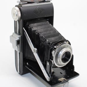 May include: A vintage black folding camera with a bellows mechanism, a silver lens assembly, and a top-mounted leather strap. The camera body features a textured finish, a silver winding knob, and a metal support arm extending to the front lens.