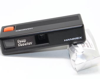 Pode incluir: Uma câmera preta Hanimex Snap Shooter com um botão laranja e uma pulseira preta. A câmera está ao lado de um pacote de filme prateado. A câmera tem as palavras "Snap Shooter" impressas.