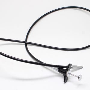 May include: A black camera shutter release cable featuring a flexible cord and a metallic silver plunger mechanism with a black plastic grip. The device includes a threaded metal tip at the end of the cable, designed for manual remote photography.