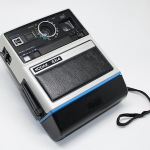 Puede incluir: Una cámara instantánea Kodak EK4 vintage. La cámara tiene un cuerpo plateado y negro con una franja azul. La lente está centrada y la cámara tiene un agarre de cuero negro. Se adjunta una correa de muñeca negra.