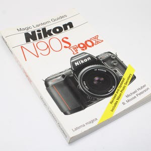 Könnte beinhalten: Ein Taschenbuch Magic Lantern Guide für Nikon N90s und F90x Kameras. Das weiße Cover zeigt eine schwarze Nikon Kamera mit Objektiv, roten und schwarzen Text sowie ein gelbes diagonales Banner mit der Aufschrift Revised and Updated! Includes Nikon.