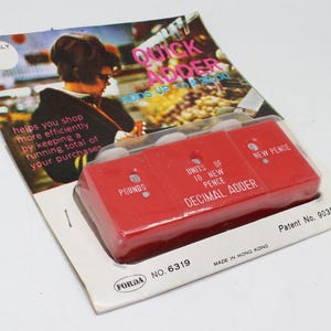 May include: A vintage red decimal adder in its original packaging. The device is labeled with "Pounds," "Units of 10 New Pence," and "New Pence." The packaging includes the text "Quick Adder" and "Helps you shop more efficiently."