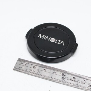 May include: A black Minolta camera lens cap with the brand name in white. A stainless steel ruler is next to the cap, showing inches and centimeters.