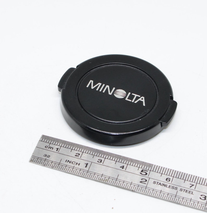 May include: A black Minolta camera lens cap with the brand name in white. A stainless steel ruler is next to the cap, showing inches and centimeters.