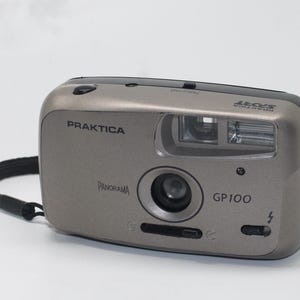 Op de afbeelding: Een zilveren Praktica GP100 Panorama camera met een zwarte polsband. De camera heeft een lens, flitser en diverse knoppen. Het woord "PRAKTICA" staat op de voorkant.
