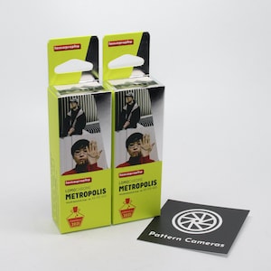May include: Two packs of LomoChrome Metropolis color negative film, each with a green and silver box featuring a photo of a person holding their hand up. The text on the box reads "LomoChrome Metropolis Color Negative Film 35mm XR 100-400 Formula 2021."