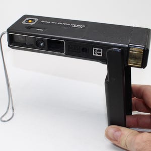 Könnte beinhalten: Eine schwarze Kodak Tele-EKTRALITE 600 Kamera mit rechteckigem Gehäuse, gelbem Auslöser, Objektiv, Blitzgerät auf der rechten Seite und silberner Handschlaufe. Die Kamera verfügt über einen klappbaren Griff und das Kodak-Logo auf der Vorderseite.