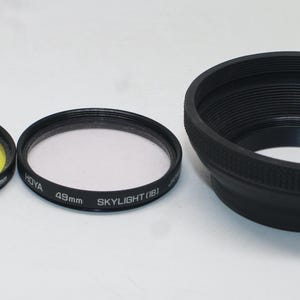 Op de afbeelding: Drie camera-accessoires: een gele kleurspotfilter, een helder skylightfilter en een zwarte zonnekap. De filters hebben een diameter van 49 mm. De zonnekap is zwart en heeft een geribbeld ontwerp.