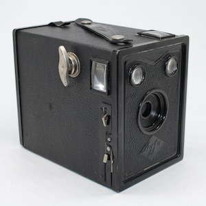 Puede incluir: Una cámara Agfa de caja negra vintage con un asa de cuero y detalles plateados. La cámara cuenta con una lente, un visor y una llave de cuerda. El logotipo de Agfa es visible en la parte frontal de la cámara.