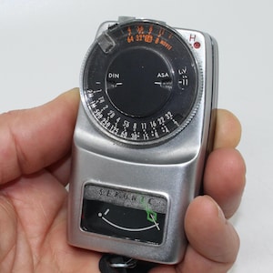 Puede incluir: Un fotómetro Sekonic plateado con diales negros y una cubierta de plástico transparente. El medidor tiene varias configuraciones para ASA, DIN y modo película. El dispositivo se sostiene en una mano.
