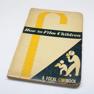 May include: A vintage book titled "How to Film Children." The cover is beige with a large yellow "F" design and the title in black. A dark blue triangle features illustrations of a person filming a child. The book is part of "A Focal Cinebook" series.