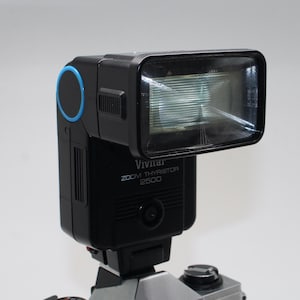 May include: A black Vivitar Zoom Thyristor 2500 flash unit with a rectangular flash head. The flash has a blue accent and is mounted on a camera. The text "Vivitar ZOOM THYRISTOR 2500" is printed on the front.