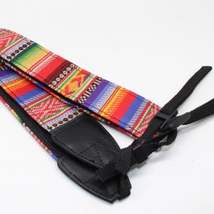 May include: A camera strap featuring a vibrant, multi-coloured woven pattern with stripes of red, blue, yellow, and green. The strap includes black leather end tabs and adjustable black nylon webbing connectors for secure attachment to a camera body.