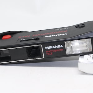 May include: Black Miranda Photosport Tele camera with a red button and a black wrist strap. The camera has a built-in flash and the words "MIRANDA PHOTOSPORT TELE" are printed on the front in red and white. A roll of film is next to the camera.
