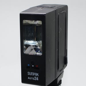 May include: A black Sunpak Auto 24 camera flash unit. The flash has a rectangular shape with a clear lens and a control panel with red and blue indicators. The text "SUNPAK AUTO 24" is printed on the front.