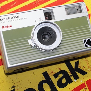 May include: A silver and green Kodak Ektar H35N half frame camera with a black lens and a silver lens ring. The camera has a black Kodak logo on the side. The text "EKTAR H35N HALF FRAME CAMERA" and "Kodak" are printed on the front of the camera.