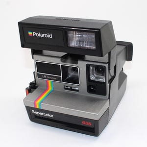 May include: A black and grey Polaroid Supercolor 635 instant camera featuring a prominent rainbow stripe graphic on the front. The device includes a built-in flash unit, a lens, a viewfinder, and the text Polaroid, LM Program, and 635.
